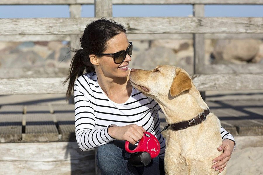 Ver imagen 5 de 6: Mujer con gafas de sol sujetando una flexi Classic roja junto a un labrador claro apoyado en un muro